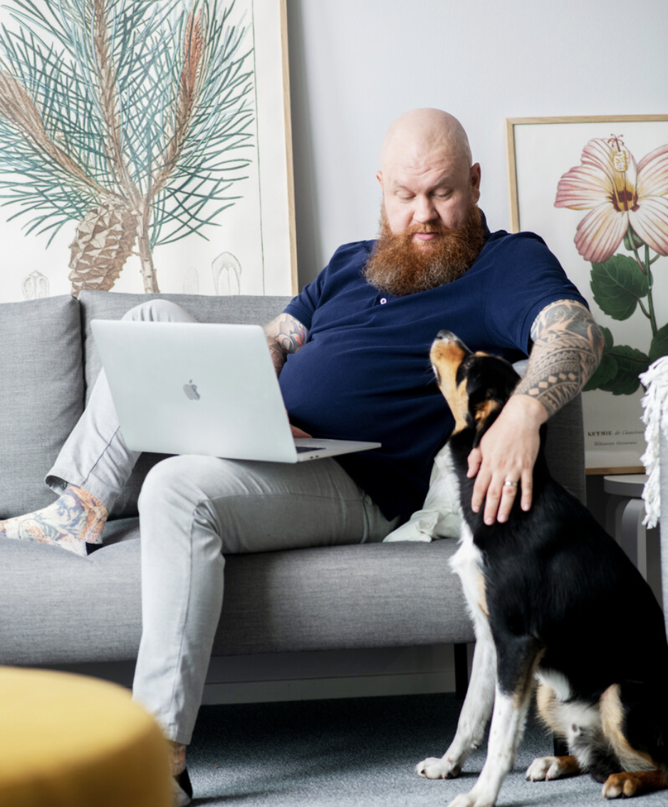 Mann som sitter i en sofa med laptopen og koser med hunden sin
