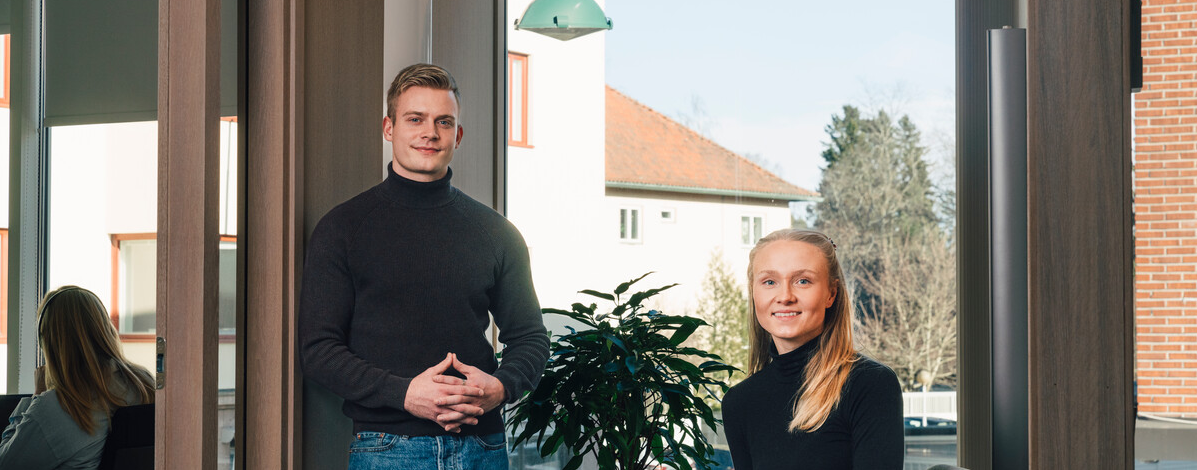 Ung mann og kvinne som står og smiler mot kamera i et kontorlandskap. hun sitter til høyre for han i en stol, mens hans står til venstre. Begge har på seg en svart turtleneck og blå jeans.