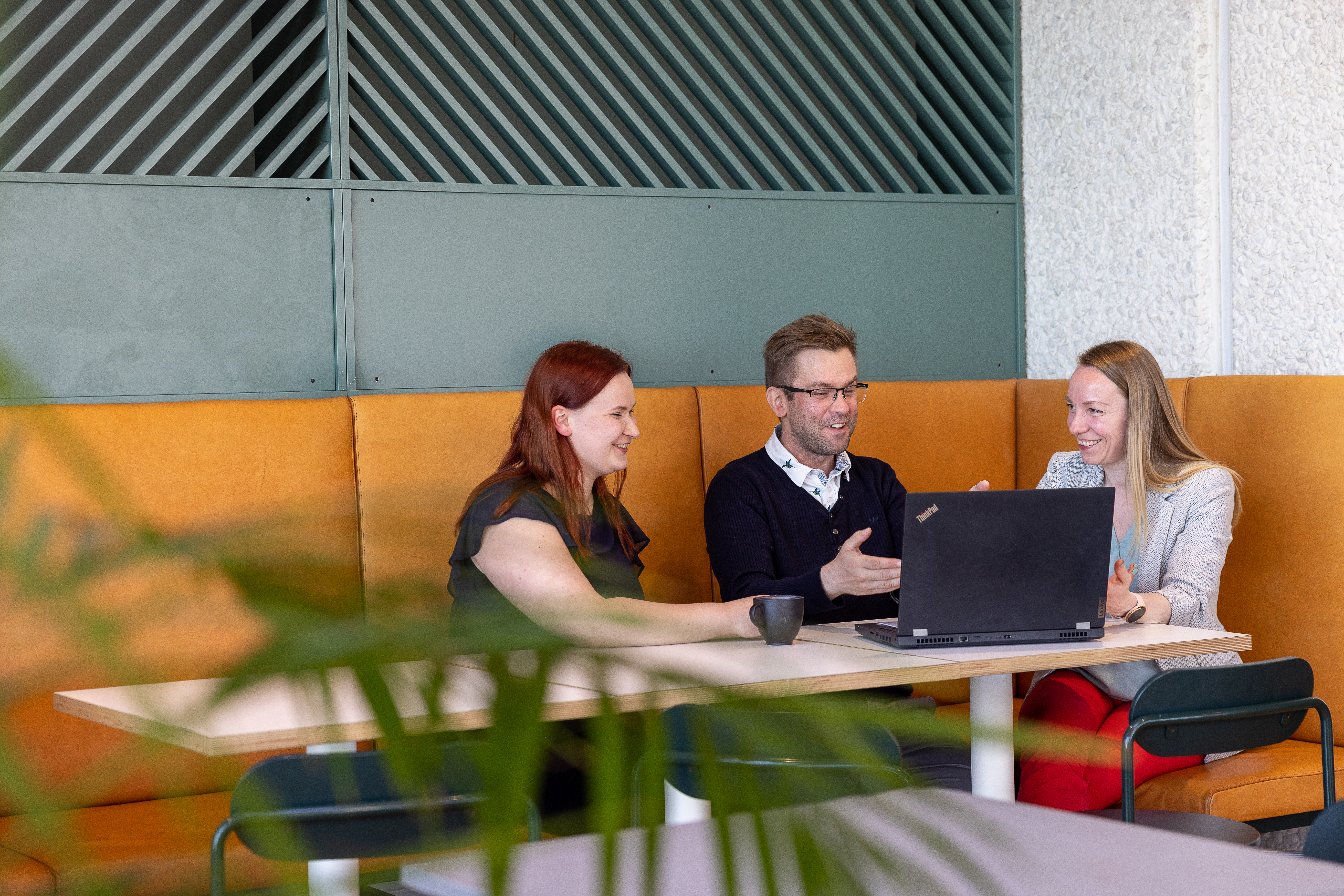 Tre kolleger kolleger i en oransje sofa med grønn vegg og et par kaffekopper